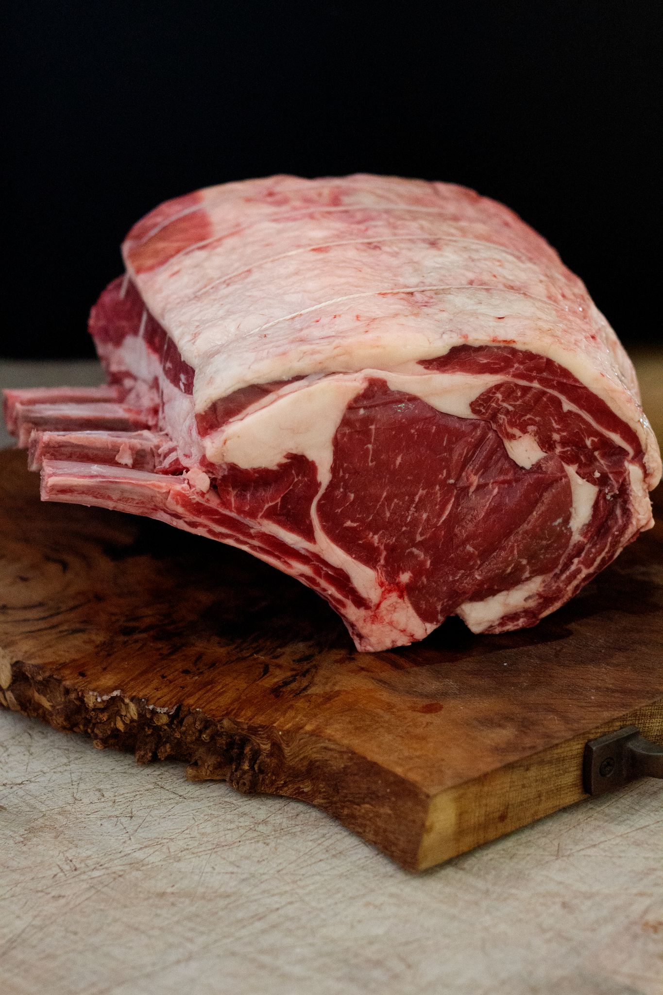 A large joint of beef rib, placed on a wooden chopping board, against a black background