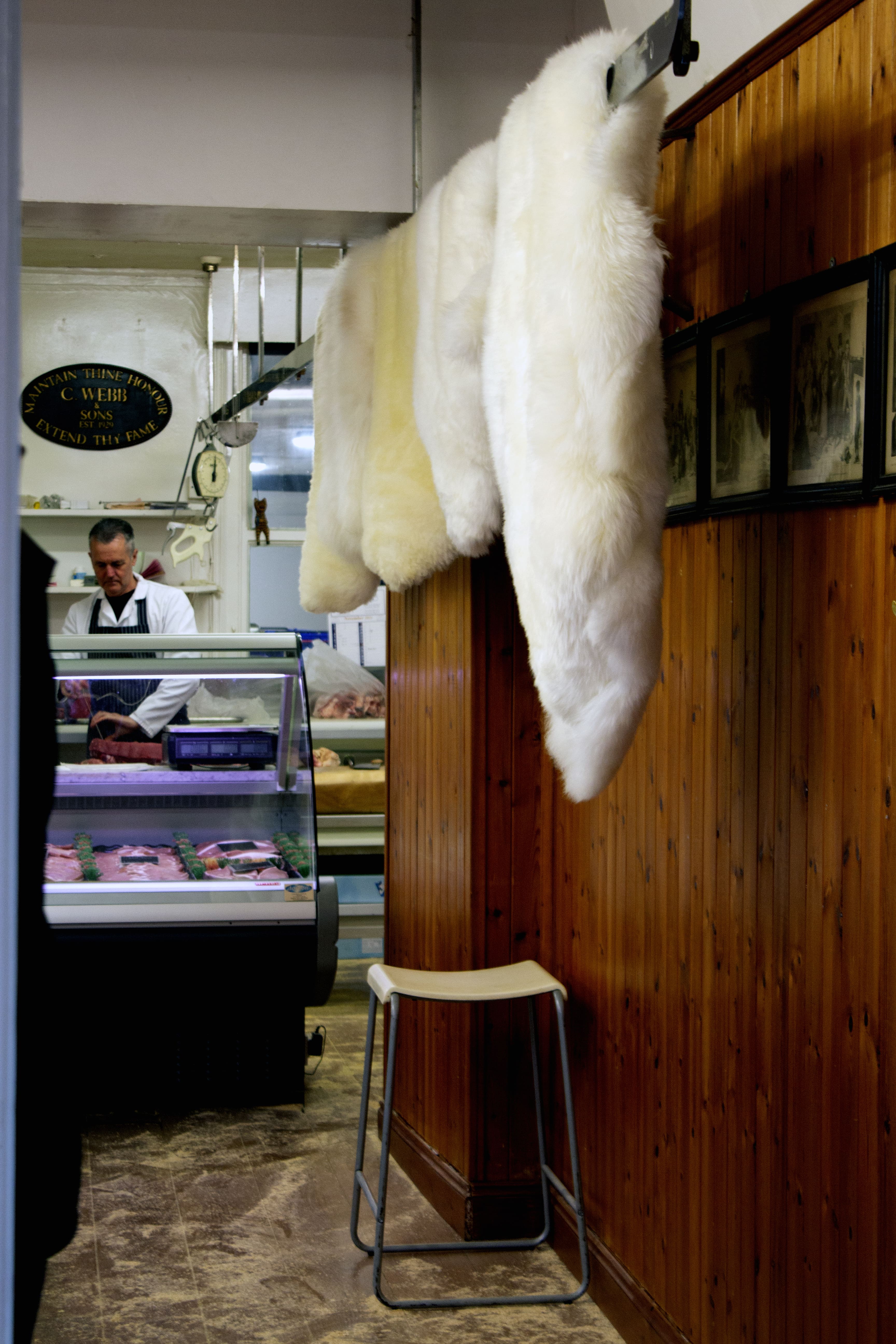 An image of inside the C Webb & Sons Butchers Shop, showing some of the counter, a butcher in the background and sheep skin rugs for sale.