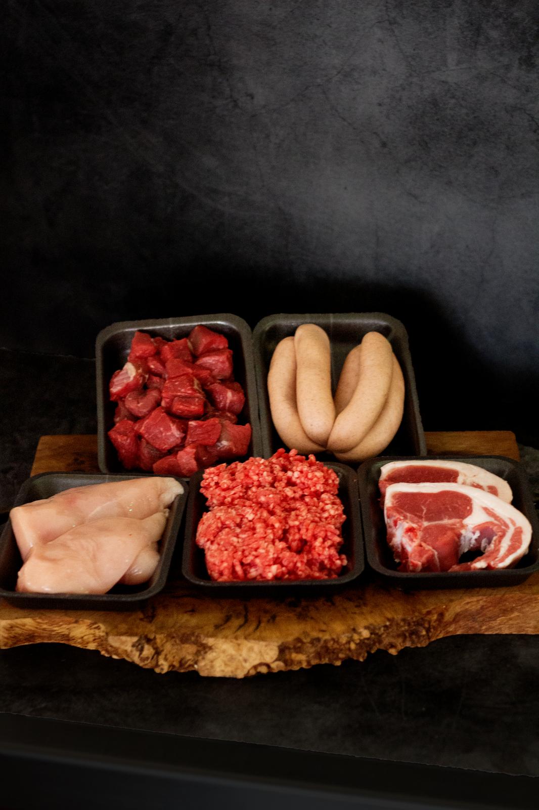 An image showing five trays containing; pork sausages, braising steak, chicken fillets, beef mince and chops. All on a wooden chopping board with a black background.