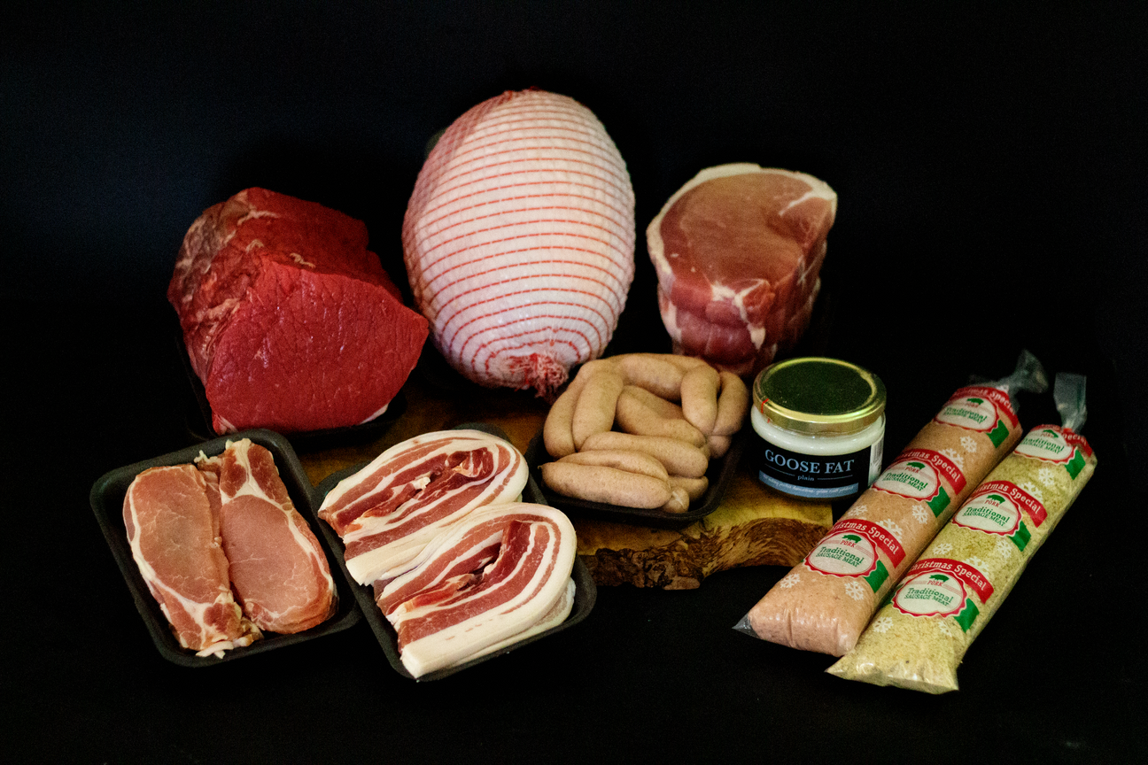 A Christmas meat hamper on a black background, featuring rolled turkey breast, beef roasting joint, gammon joint, back bacon, streaky bacon, chipolata sausages, sausage meat, stuffing mixes, and a jar of plain goose fat.